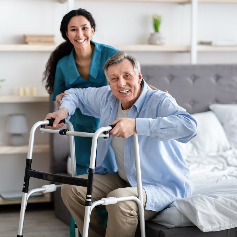 female-nurse-helping-elderly-male-with-walking-frame-stand-up-from-bed-at-home-professional-care.jpg